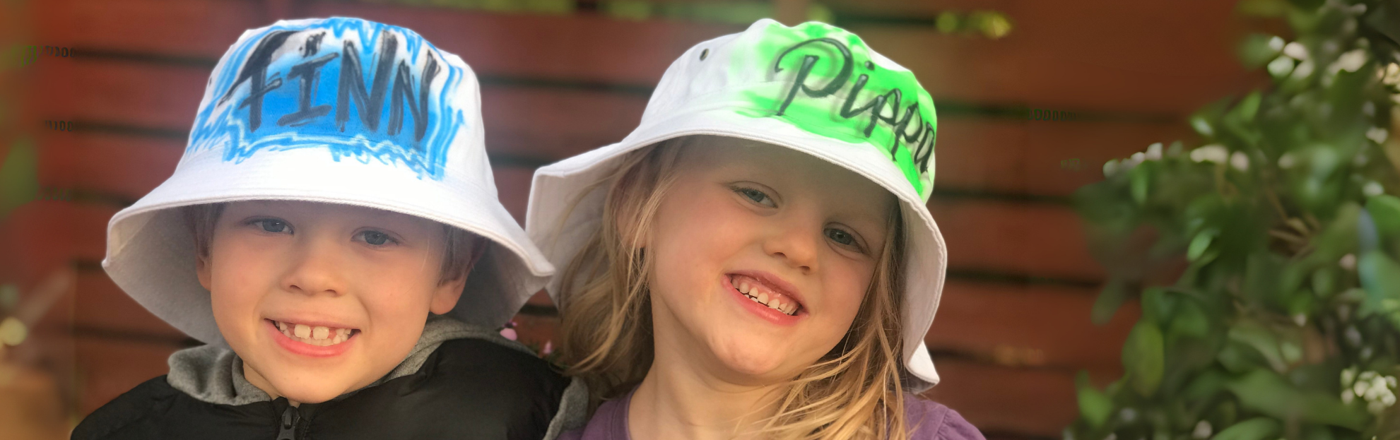 Two children wearing bucket hats with airbrushed names written on them, standing outdoors.
