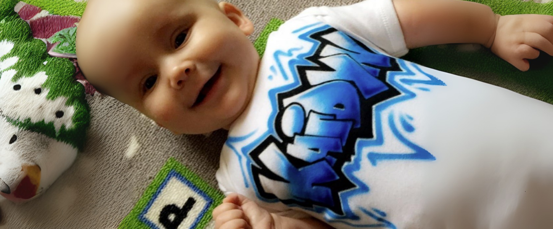Baby wearing a white onesie with blue graffiti name graphics, lying on a carpeted floor.