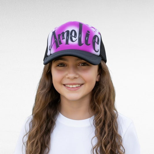Young girl wearing a cap with 'Amelie' airbrushed on it with a purple design against a white background
