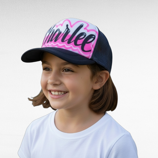 Child wearing a black trucker hat with 'Charlee' airbrushed with a pink design on a plain background