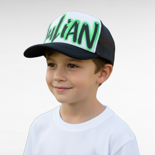 Child wearing a black and white trucker cap with green airbrush text Julian on a plain background